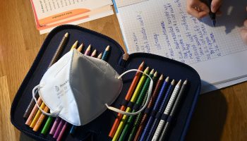 A protective face mask lies on a pencil case as a girl does her school work at a writing desk.