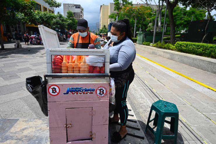 Two people wearing masks stand working at an ice cream cart the shows stickers on the side with the Bitcoin icon crossed out.