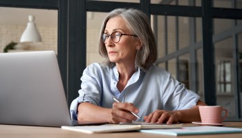 Serious mature older adult woman watching training webinar on laptop working from home or in office. 60s middle aged businesswoman taking notes while using computer technology sitting at table.