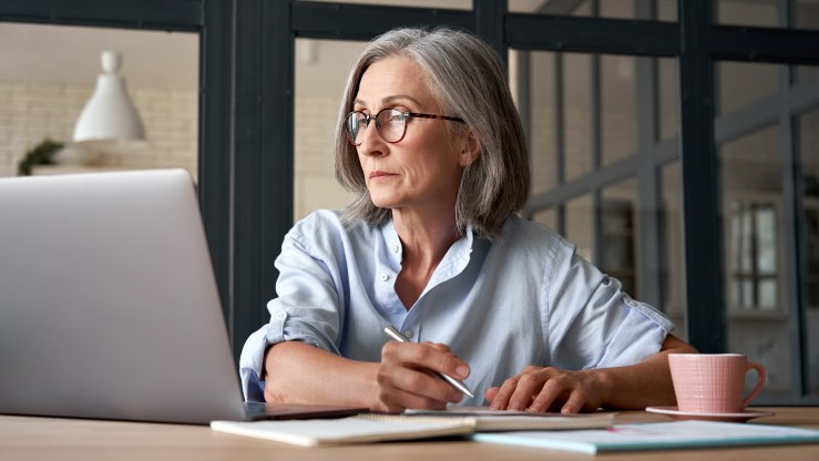 Serious mature older adult woman watching training webinar on laptop working from home or in office. 60s middle aged businesswoman taking notes while using computer technology sitting at table.