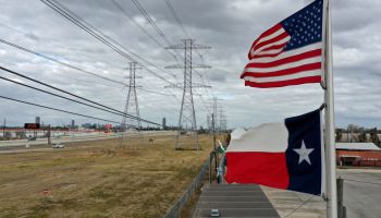 The American flag flies above the Texas flag, with power lines in the background.