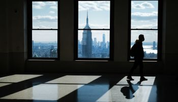 A figure in silhouette walks through an empty office building with a view of Manhattan outside the the tall windows.