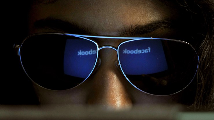 The Facebook logo is reflected in a girl's sunglasses as she looks at her screen.