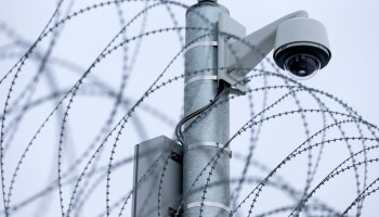 A surveillance camera located on a prison perimeter sits upon a pole above barbed wire.