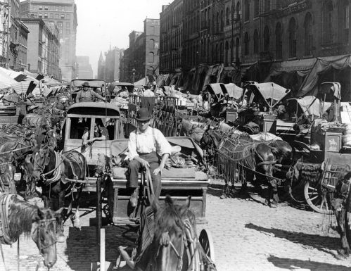 A crowded street in Chicago can be seen bustling with horse drawn carriages. A man on a carriage rides towards the photographer.