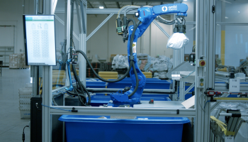 A blue Ambi robot uses suction to grip a package midair as it lifts it out of a blue bin.
