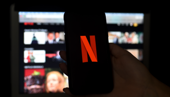 A hand holds an iPhone with the Red "N" Netflix logo. In the background, there is a computer screen with reels of still shots from Netflix movies and shows.