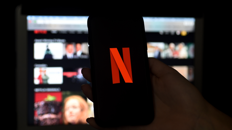 A hand holds an iPhone with the Red "N" Netflix logo. In the background, there is a computer screen with reels of still shots from Netflix movies and shows.