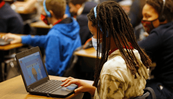 A student sits masked in a classroom full of other masked children. Each student completes a task on their computer while wearing a set of navy headphones.