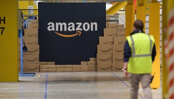 An Amazon employee walks toward a stack of packages and a large sign displaying the company's name and logo.