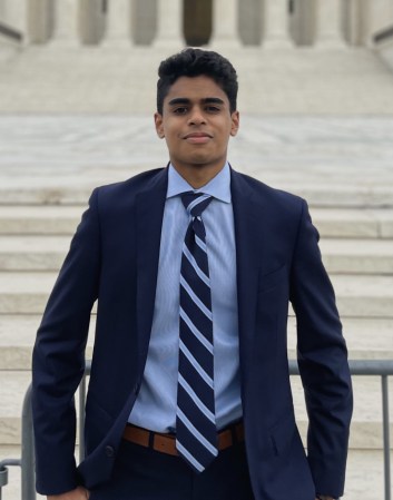 Zamaan Qureshi stands wearing a blue blazer and striped tie in front of a flight of marble steps.
