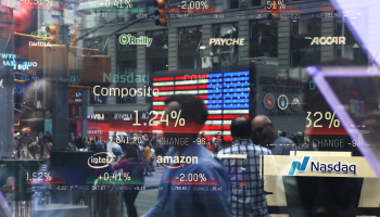 A man in a collared shirt walks past a window of screens showing companies and their stocks. These include many tech companies like Amazon and Intel.