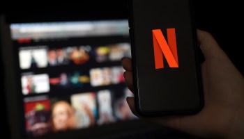 A person hold a phone with the Netflix logo in front of a laptop with the Netflix home screen shown.
