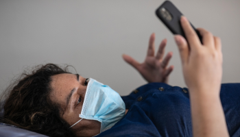 A woman wearing a face mask and laying on her bed conducts a Zoom call with a doctor.