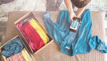 A woman takes a picture of a blue denim shirt with a smartphone. Boxes of clothes are next to the shirt.