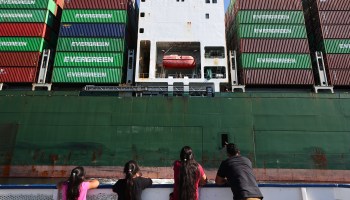 People ride on a tour boat beneath cargo containers stacked on a container ship at the Port of Los Angeles.