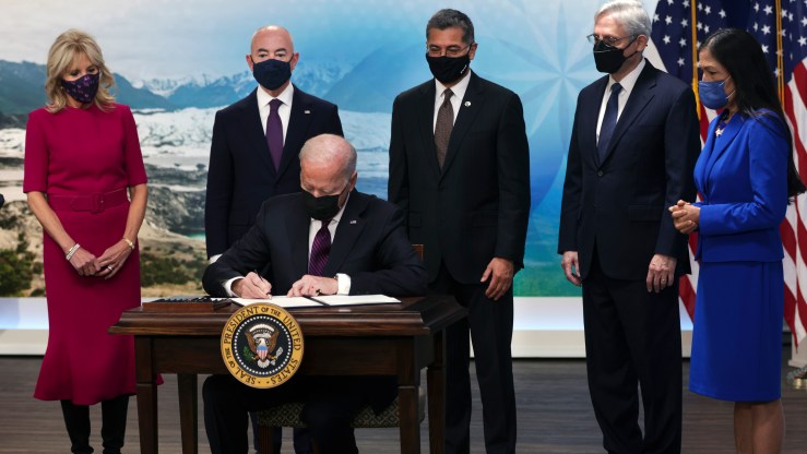 President Joe Biden sits at a table as he signs an executive order to help improve public safety and justice reform for Native American communities. People wearing face masks stand behind him.