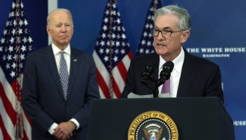 Federal Reserve Board Chair Jerome Powell, right, speaks at a lectern with the presidential seal as President Joe Biden listens. There are three American flags in stands behind them.