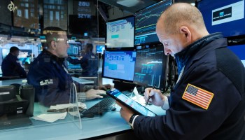 Traders sit at desks at desks on the floor of the New York Stock Exchange, with a clear partition separating them. One works on a tablet, the other at a computer with several monitors.