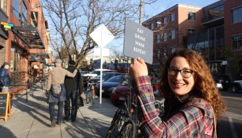 Megan Bucholz, founder of Local Table Tours, stands on a city street holding a sign that reads "Eat Drink Walk Repeat."