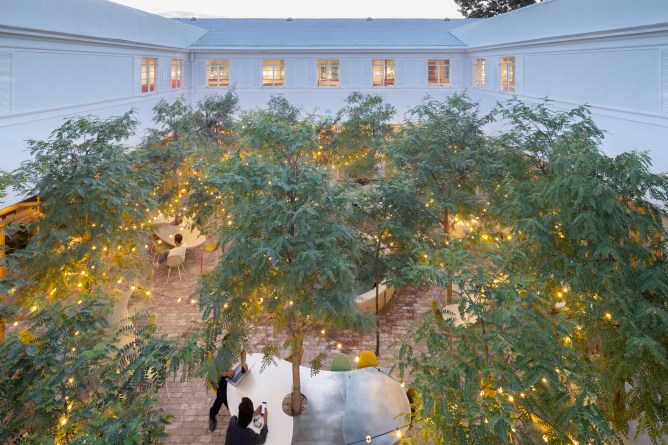 People work at computers in a large brick patio at Second Home Hollywood, where trees grow through tabletops. A string of lights run through the trees.