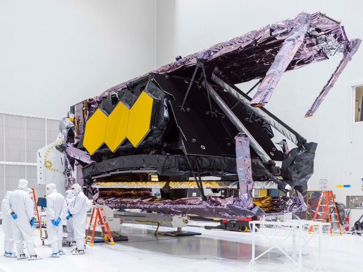 The James Webb Space Telescope, which is silver, black and has three hexagonal yellow plates on the side, is folded horizontally after its removal from the packing container.