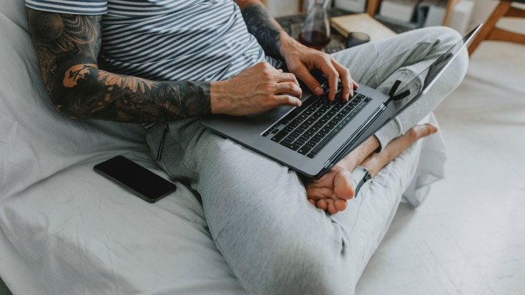 A man works on his laptop in a striped T-shirt and sweatpants.
