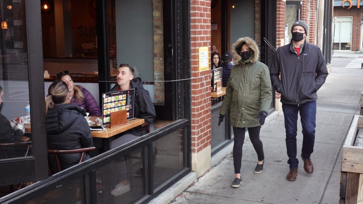 Patrons sit at an open-air restaurant as people pass by on the sidewalk.