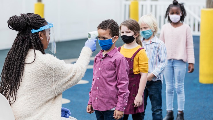 A nurse with a face shield takes the temperatures of masked schoolchildren.