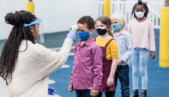 A nurse with a face shield takes the temperatures of masked schoolchildren.