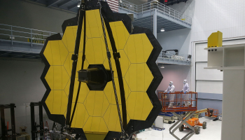 The James Webb Space Telescope, which is bright yellow and has a honeycomb like structure, stands upright in a large lab. NASA employees in paper suits operate on the telescope.