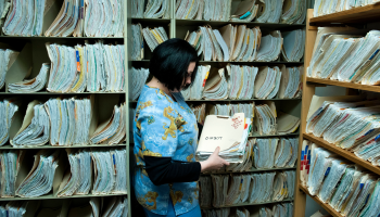 A woman wearing blue scrubs holds a manila folder overflowing with papers and stands in front of floor-to-ceiling shelves of full manila folders.