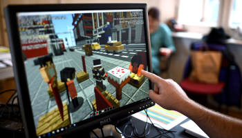 A hand points at a Dell monitor with a Minecraft display. The screen includes a cityscape in Minecraft including taxis and avatars.