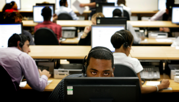 Several workers sit at computers with headsets. One employee in the center can be seen facing towards the camera with his forehead and eyes above his desktop.