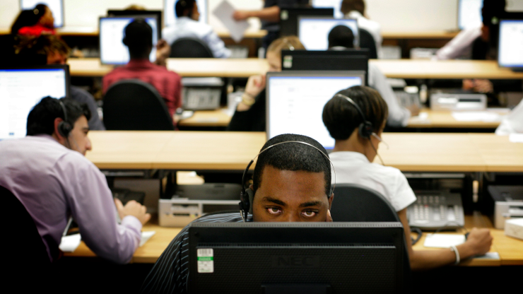 Several workers sit at computers with headsets. One employee in the center can be seen facing towards the camera with his forehead and eyes above his desktop.