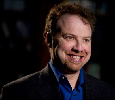 Adam Riess smiles wearing a blue collared shirt and a black suit jacket.