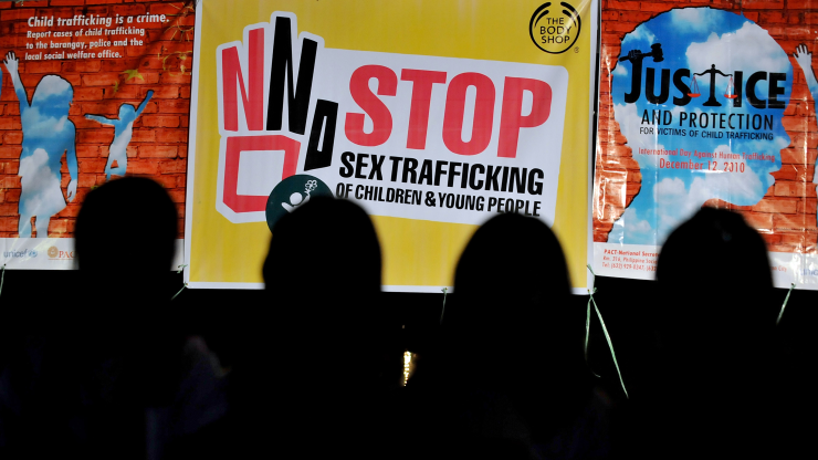 Four people are backlit by three posters. The central one is yellow, pink, white and black and it says, "Stop sex trafficking of children and young people."