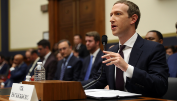 Facebook cofounder Mark Zuckerberg speaks into a microphone at a desk as he testifies. Behind him, there are a couple of rows of people in suits listening.