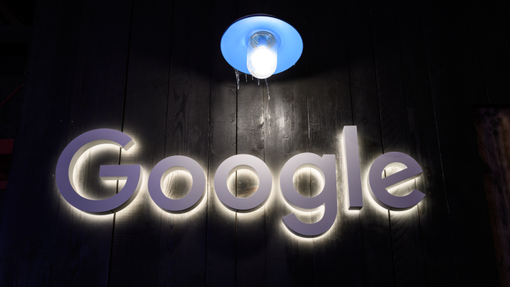 A white sign that reads "Google" hangs on a dark wall and is illuminated by a single lightbulb.