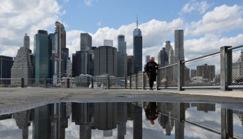 A woman wearing a face mask and the skyline of lower Manhattan are reflected in a puddle after heavy rain.