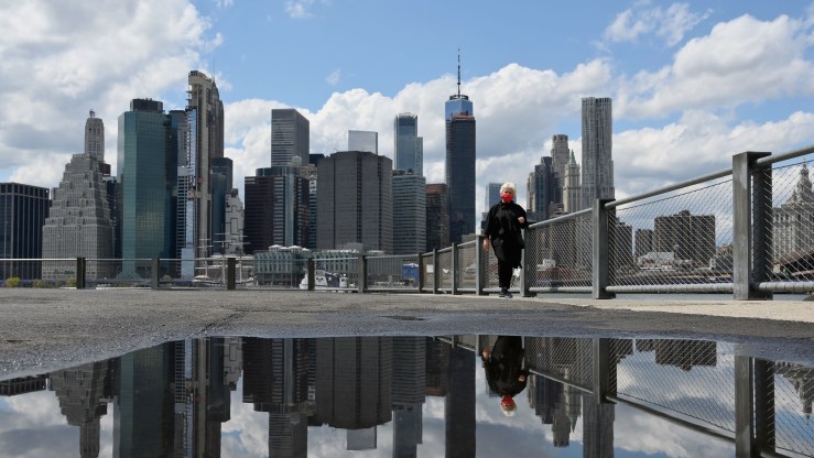 A woman wearing a face mask and the skyline of lower Manhattan are reflected in a puddle after heavy rain.