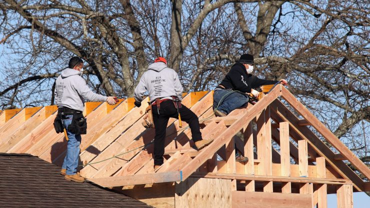 Three construction workers building a roof on top of an unfinished house