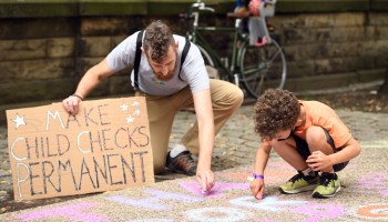 A man holding a sign that says "Make Child Checks Permanent" draws with chalk on a sidewalk next to a child.