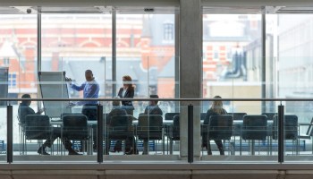 Group of six business people in a boardroom meeting. Shot at a distance from outside through the glass.