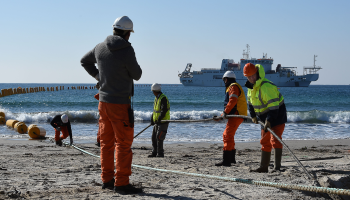 Five employees wearing helmets and neon orange and yellow gear hold a submarine cable wire and place it on the sand. In the background, there is a view of a body of water, with a boat in the distance.