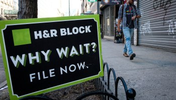 An H&R Block sign says "Why wait? File now" as a pedestrian walks in the background.