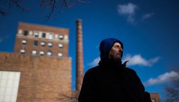 Dale Bonanno poses for a photo in front of a large brick building and chimney. Behind him, the sky is clear blue and is face is partially covered by a shadow.
