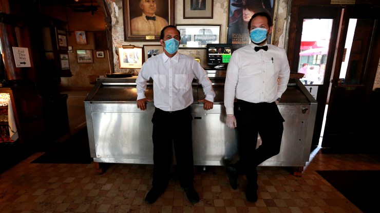 Two waiters wearing white shirts, black pants and blue face masks stand next to a bar.