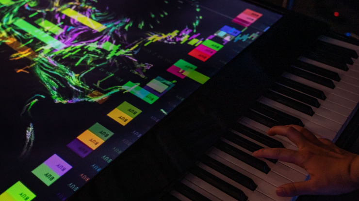 A hand plays keys on a piano with a bright colorful screen just above.