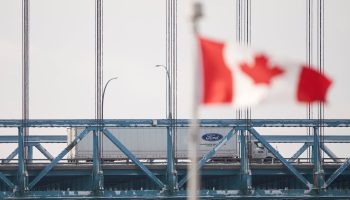 A truck drives across the Ambassador Bridge in Canada, with a Canadian flag flying in the foreground.